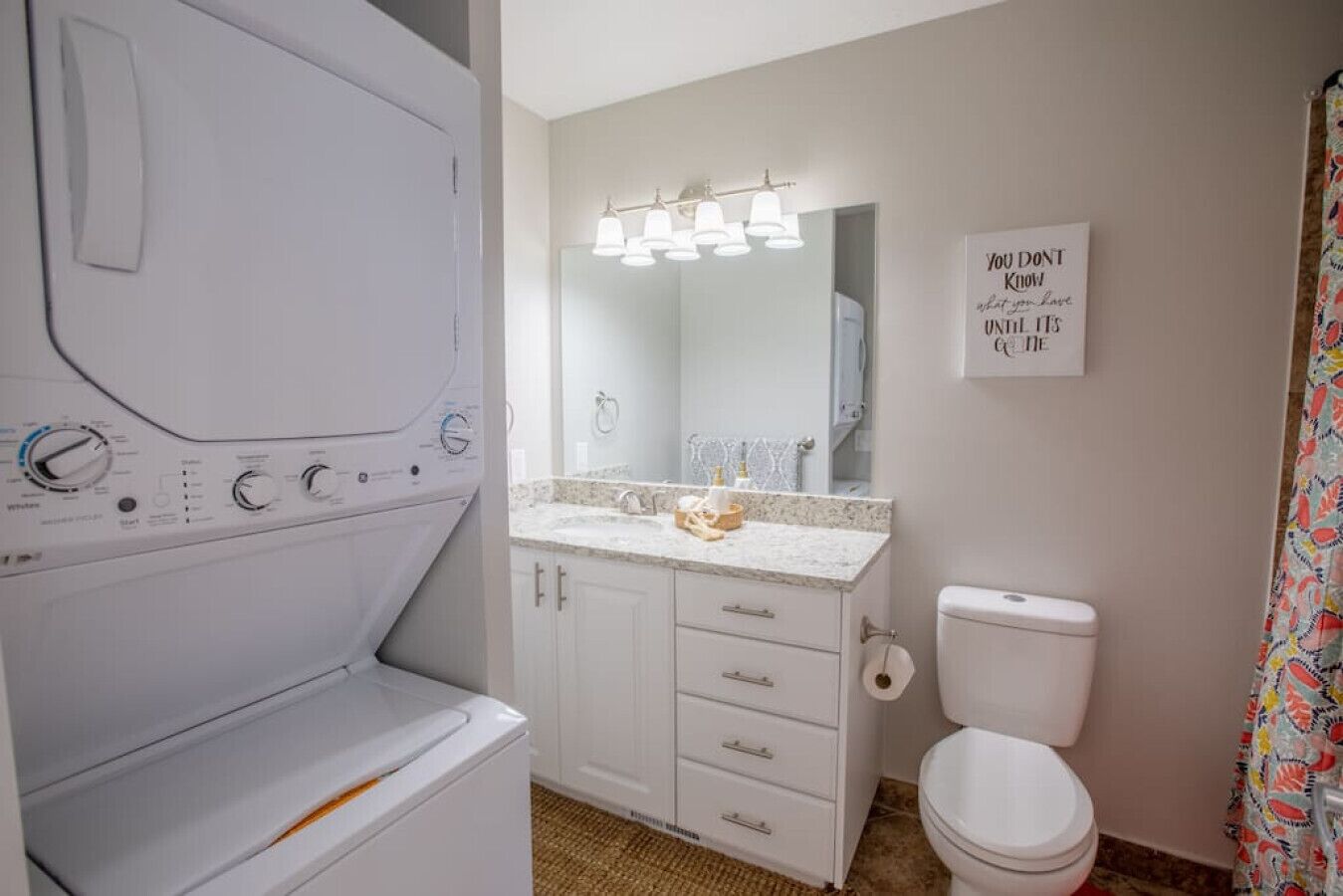 Une salle de bain équipée d’un lave-linge, d’un lavabo avec un miroir, de toilettes et d’un panneau décoratif au mur.