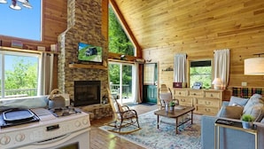 A cozy cabin interior with a stone fireplace, wooden ceiling, and large windows offering a view of the outdoors.