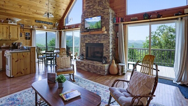 A cozy cabin interior with a stone fireplace, wooden furniture, and large windows offering scenic mountain views.