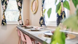 Zona pranzo con un tavolo lungo, sedie rosa e tende floreali.