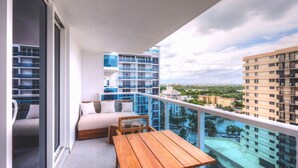 A balcony with a sofa and a wooden table, overlooking a cityscape with modern buildings and a pool. - Oceanview Private Condo at 1 Hotel & Homes -1211 (Miami Beach)