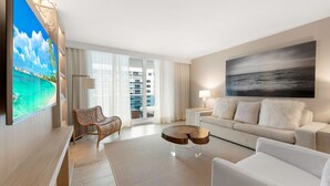 A modern hotel room with a large TV, a sofa, a chair, a coffee table, and a view of the city through large windows. - Oceanview Private Condo at 1 Hotel & Homes -1211 (Miami Beach)