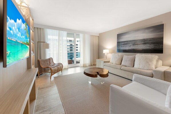 Ein modernes Hotelzimmer mit großem Fernseher, einer Couch, einem Sessel, einem Couchtisch und Blick auf die Stadt durch große Fenster.