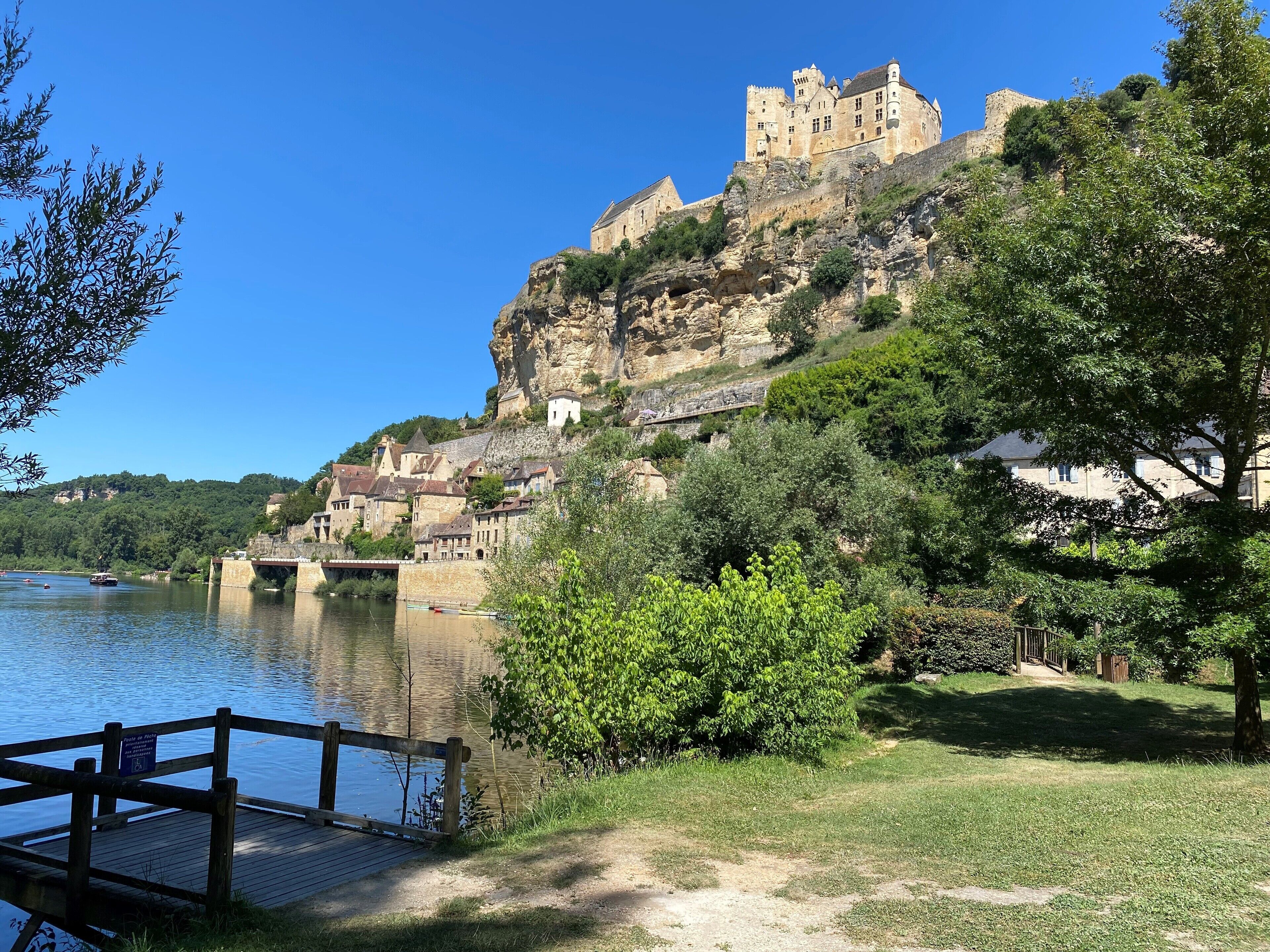 A historic castle on a hill overlooking a river, with a wooden dock and lush greenery.