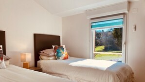 A hotel room with two beds, a nightstand with a lamp, a window with a view of a garden, and a wall-mounted thermostat.