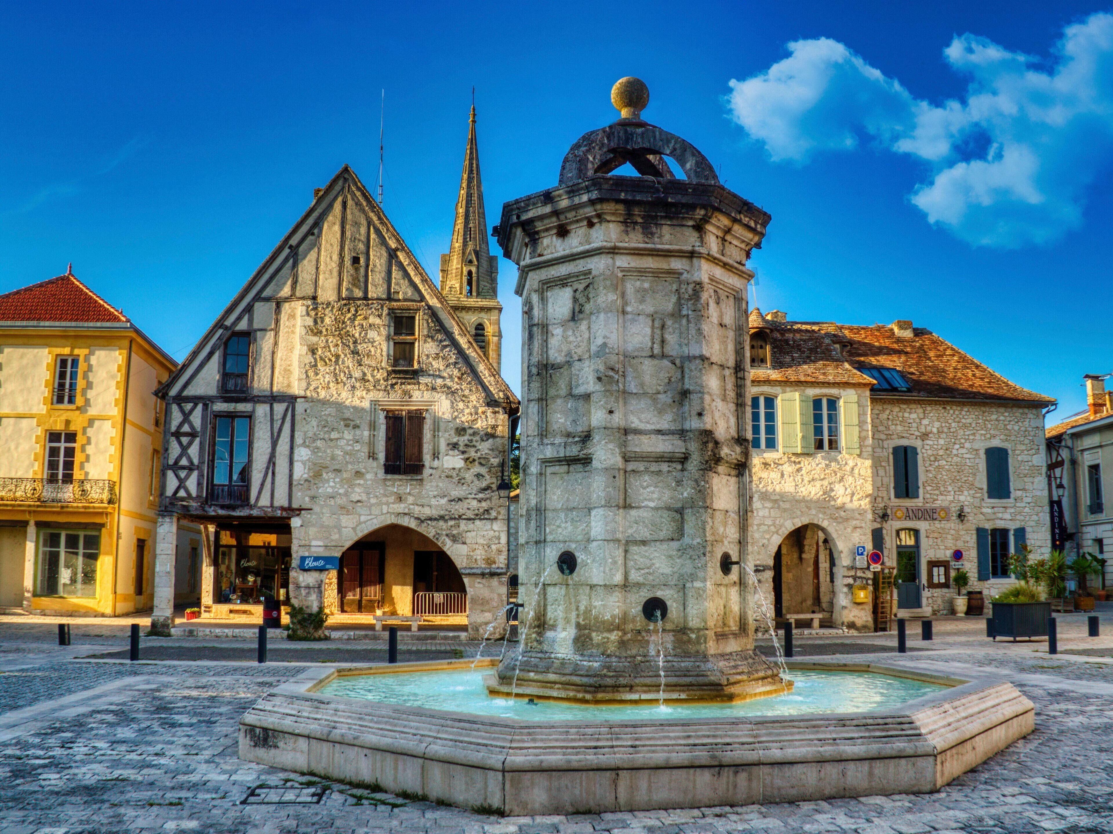 Une fontaine historique en pierre, surmontée d’un dôme, entourée d’un pavage de pierres, se dresse devant un bâtiment européen traditionnel doté d’un toit en pente et de fenêtres en arc de cercle.
