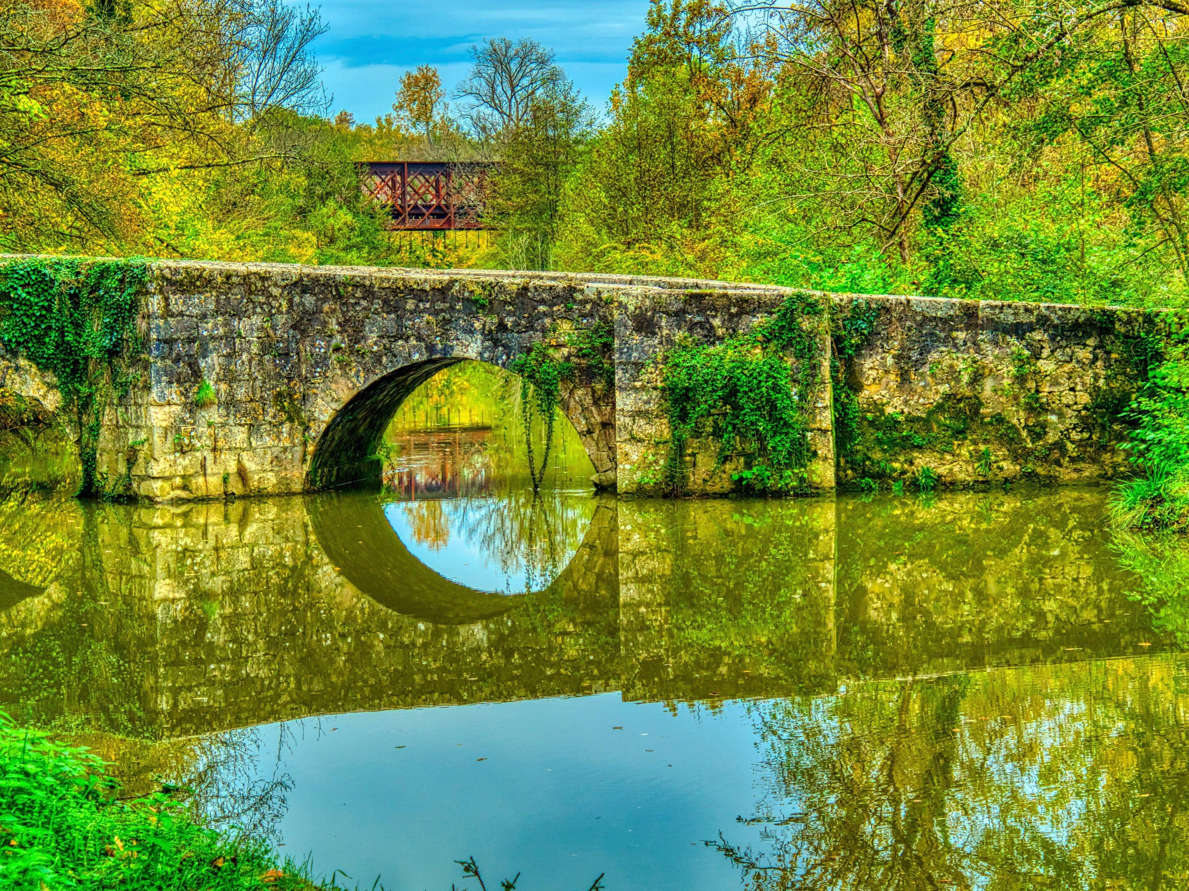 A stone bridge over a calm river with a reflection of the bridge and trees.