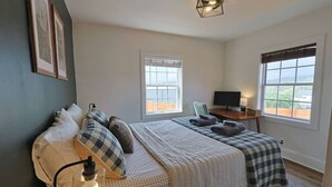 Un dormitorio con una cama, dos ventanas, un escritorio con computadora y una lámpara.
