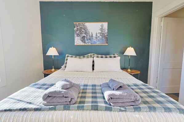 A neatly made bed with a checkered bedspread and two folded towels, flanked by bedside lamps, in a room with a green wall and a framed picture above the bed.