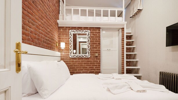 A hotel room with a brick wall, a white bed with white linens, a white-framed mirror, a white wardrobe, and a TV mounted on the wall. - White Palace Old City Boutique Hotel (Istanbul)