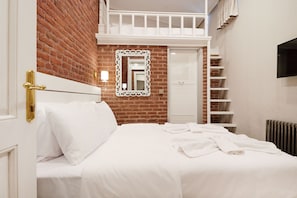 A hotel room with a brick wall, a white bed with white linens, a white-framed mirror, a white wardrobe, and a TV mounted on the wall. - White Palace Old City Boutique Hotel (Istanbul)