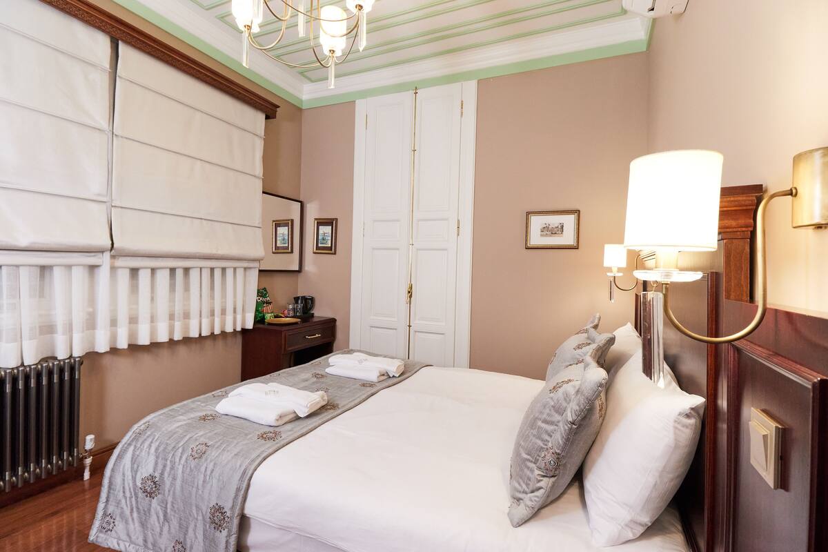 Ein Hotelzimmer mit einem großen Bett, einem Nachttisch mit Lampe, einem Heizkörper und einem Fenster mit Vorhängen.