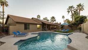 A residential swimming pool with a curved design, surrounded by a concrete deck. There are lounge chairs and a covered patio area. - Scottsdale Family Escape: Golf, Pool and Spa (Scottsdale)