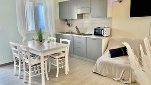 A modern kitchen with grey cabinets, a white countertop, and a microwave. There is a dining table with white chairs and a sofa with a black cushion. - Apartment 6 pers 3 mn from the beach sea view (FAVONE)