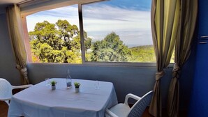 A room with a table set for two, a window with a view of trees, and curtains tied back. - Apartment 6 pers 3 mn from the beach sea view (FAVONE)