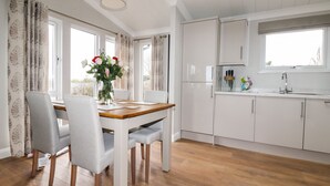 A modern kitchen with white cabinets, a wooden dining table, and a vase of flowers. - Acorn Lodge (Redruth)
