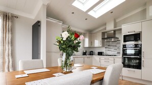A modern kitchen with white cabinets, a dining table with placemats, and a vase with flowers. - Acorn Lodge (Redruth)