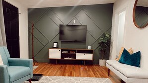 Een moderne woonkamer met een flatscreen televisie aan een geometrisch patroon muur, een blauwe fauteuil, een witte bank en een salontafel met boeken en een afstandsbediening.