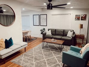 Un salon moderne avec un canapé, une table basse et un fauteuil bleu. On y trouve également un coin repas avec une table et des chaises, ainsi qu’un ventilateur de plafond.