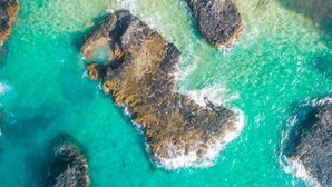 An aerial view of a coastal area with clear turquoise waters, rocky formations, and a sandy beach.