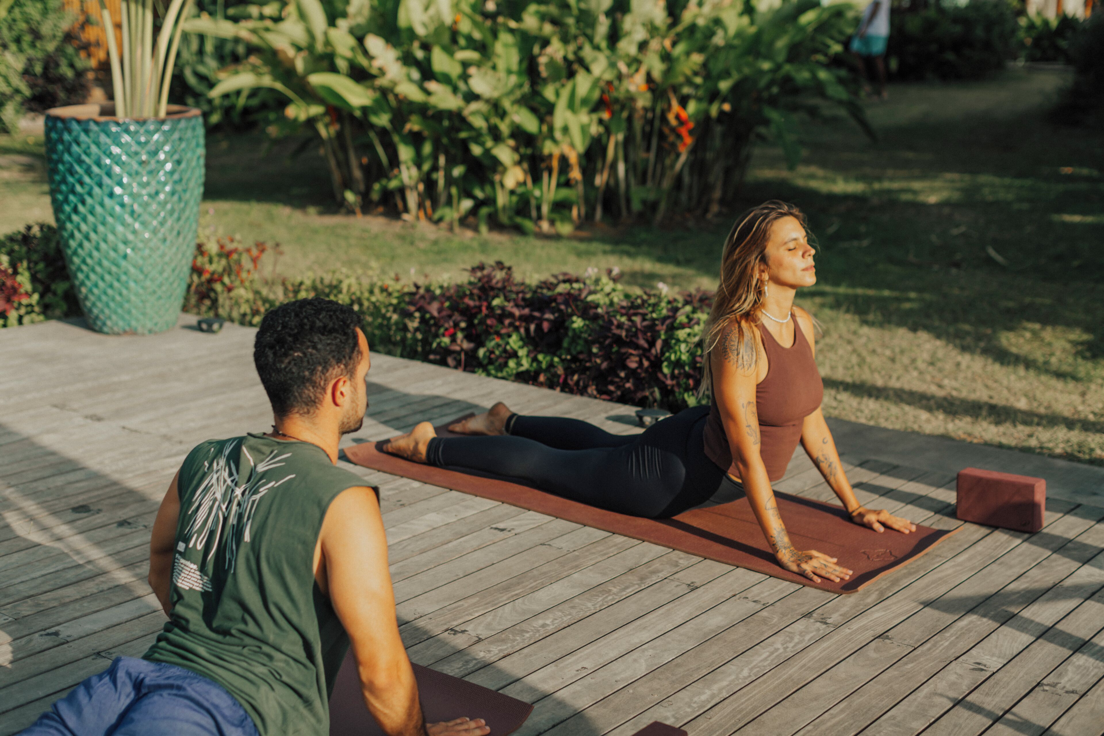 Twee personen beoefenen yoga op yogamatten in een tuin met een houten terras en potplanten.