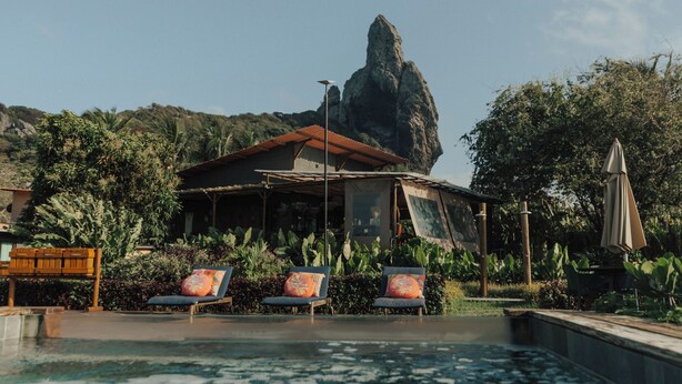 A pool with lounge chairs, a building with a red roof, and a large rock formation in the background.