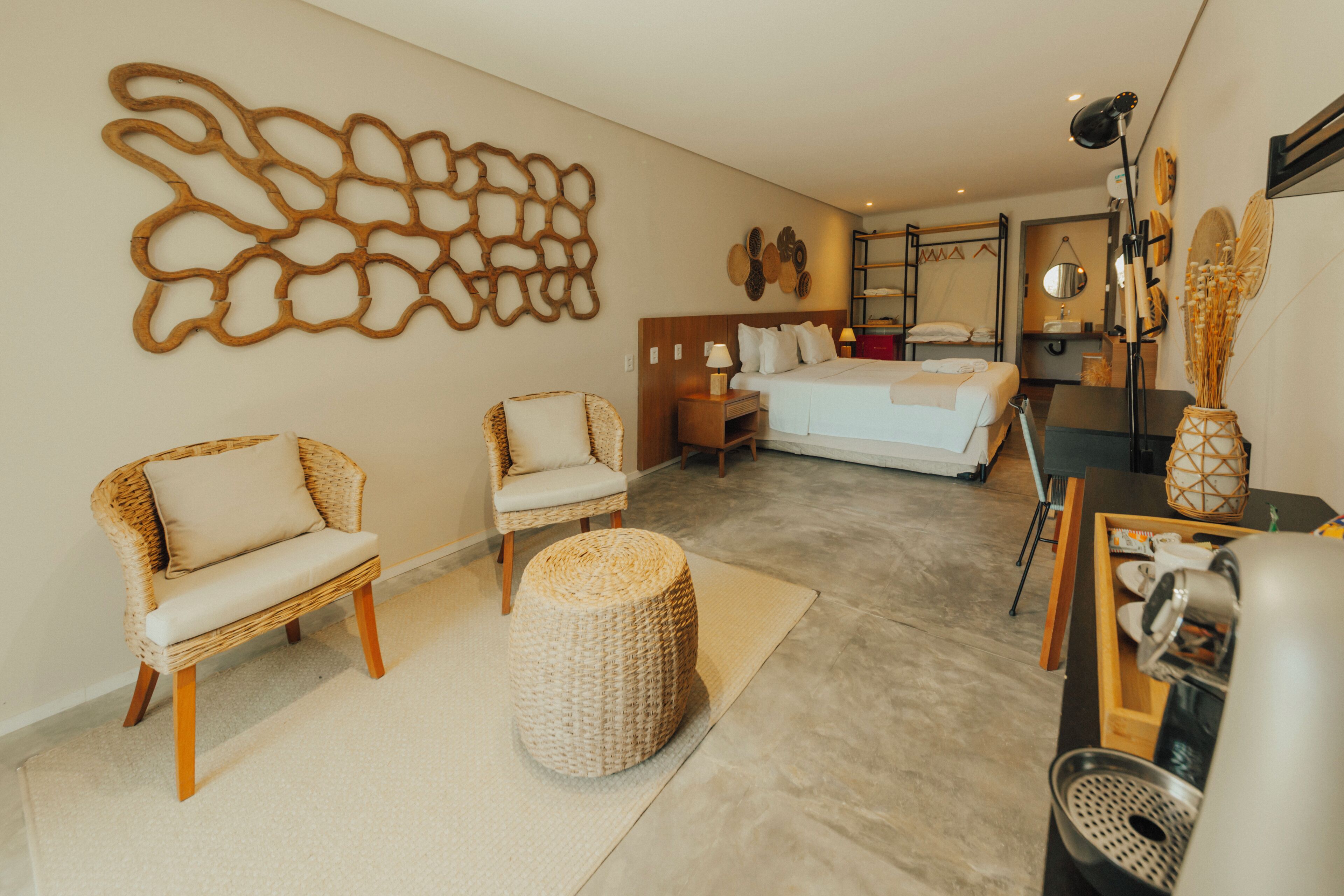 Habitación de hotel con cama, dos sillones de mimbre, un taburete redondo pequeño, cafetera y una obra de arte en la pared.