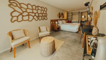 Habitación de hotel con cama, dos sillones de mimbre, un taburete redondo pequeño, cafetera y una obra de arte en la pared.
