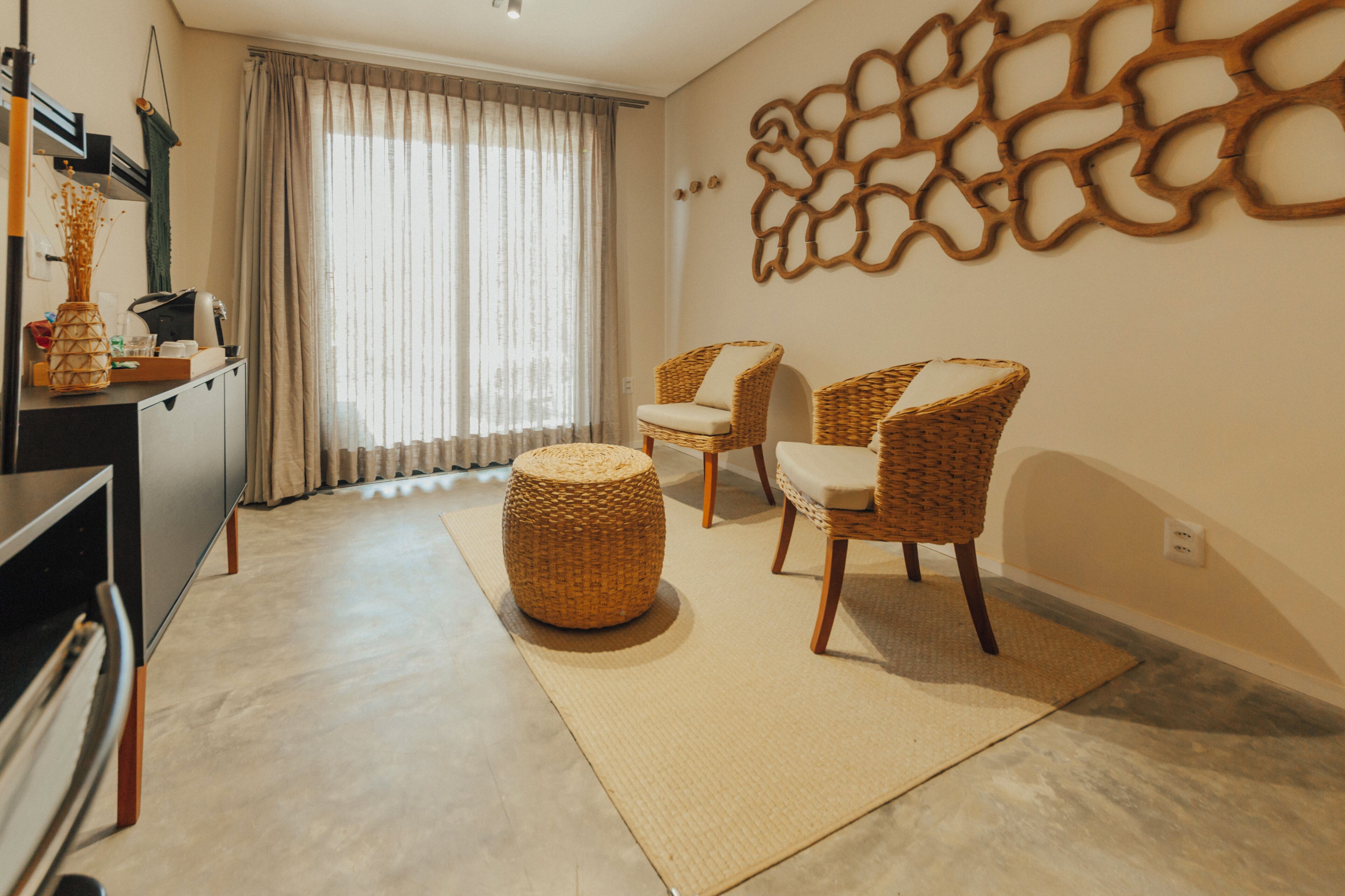 A living room with a sofa, two wicker chairs, a round ottoman, a decorative wall piece, and a window with curtains.