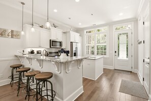 A kitchen with white cabinets, a central island with stools, and a large window with shutters.