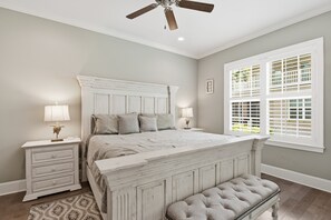 A bedroom with a large bed, a ceiling fan, a window with blinds, a bedside table with a lamp, and a bench.