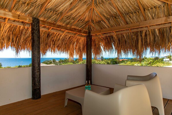 A covered outdoor seating area with a thatched roof, wooden floor, and white chairs. - Latitude Adjustment - Ocean View, Surrounded by Bonaire Nature! (Kralendijk)