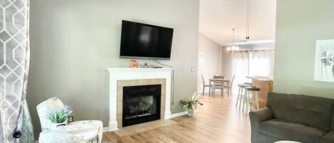 A living room with a fireplace, a television, a ceiling fan, and a dining area in the background.
