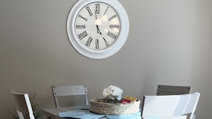 Un coin repas avec une table blanche, quatre chaises blanches, une horloge ronde fixée au mur et un panier de fleurs posé sur la table.