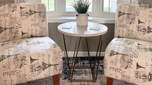 Une pièce avec deux fauteuils placés face à une grande fenêtre agrémentée de rideaux blancs, une petite table ronde sur laquelle repose un pot de fleurs et un tapis à motifs.