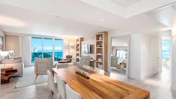 Una sala moderna con una mesa de comedor de madera, sillas blancas, un sofá y un televisor. Amplias ventanas ofrecen vistas de la ciudad.