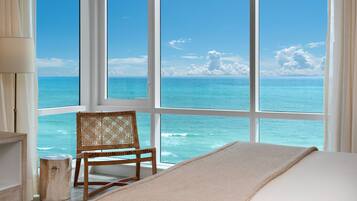 Una habitación con un amplio ventanal con vistas al océano, una silla de mimbre y una cama con una manta color beige.