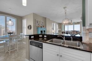 A modern kitchen with a black countertop, white cabinets, and a dishwasher. There is a large window with blinds, a chandelier, and a view of the ocean. - Beach Colony West 12A Gulf-Front 3 BD/3BA w/Beach Service & Onsite Management (Navarre Beach)
