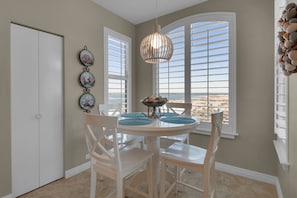 A dining area with a round table, white chairs, and a view of the beach through large windows with shutters. - Beach Colony West 12A Gulf-Front 3 BD/3BA w/Beach Service & Onsite Management (Navarre Beach)
