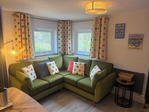 A green sectional sofa with polka dot cushions, two windows with curtains, a lamp, a poster, and a small table with a basket.