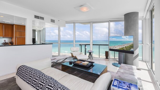 A modern living room with a large window overlooking a beach, a flat-screen TV, a sofa, and a coffee table. - Oceanfront Private Condo at The Setai -2208 (Miami Beach)