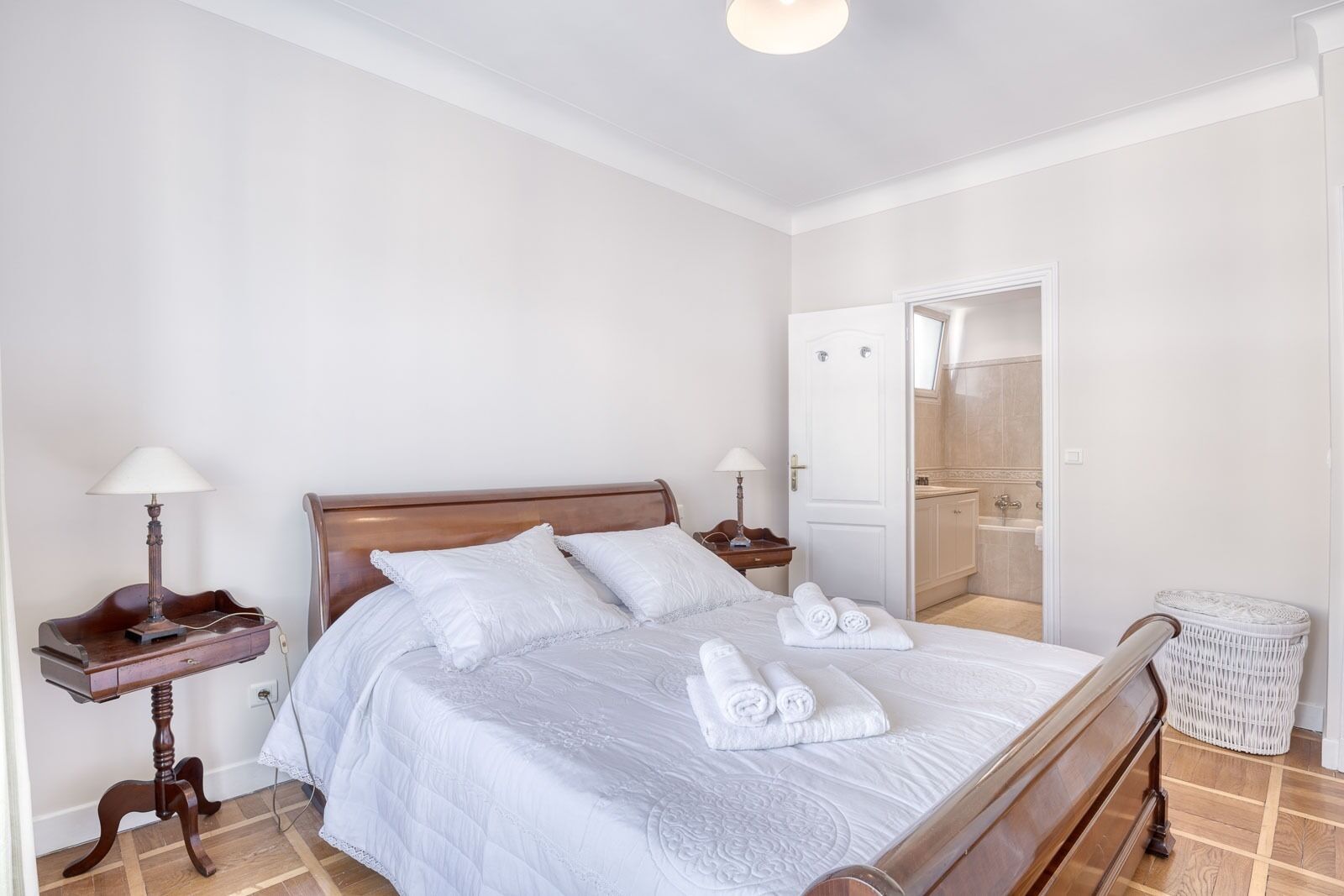 A neatly arranged bedroom with a wooden bed, white linens, a bedside table with a lamp, and a white wicker chair.