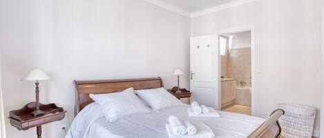 A neatly arranged bedroom with a wooden bed, white linens, a bedside table with a lamp, and a white wicker chair.