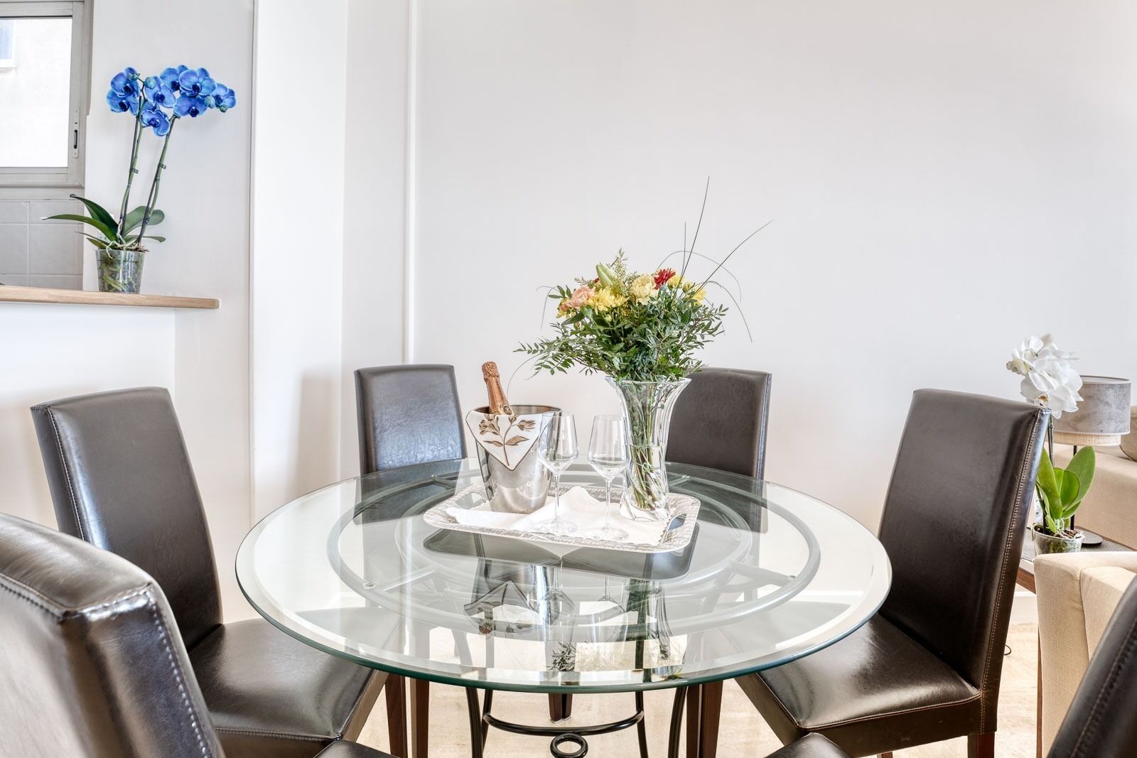 Uma mesa de vidro redonda com toalha branca, rodeada por cadeiras de couro. Um vaso com flores e uma garrafa de champanhe estão sobre a mesa.