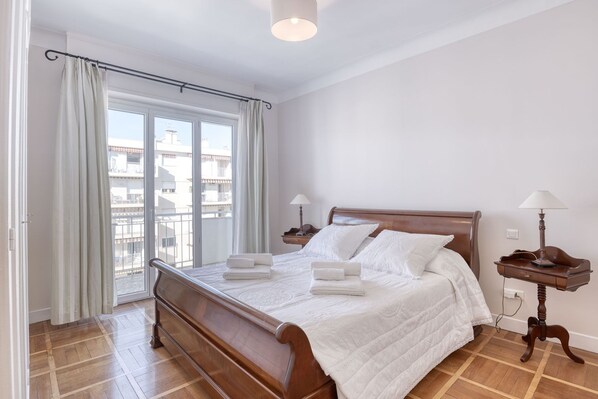Een slaapkamer met een houten bed, wit beddengoed, twee nachtkastjes met lampen en een uitzicht op gebouwen door het raam.