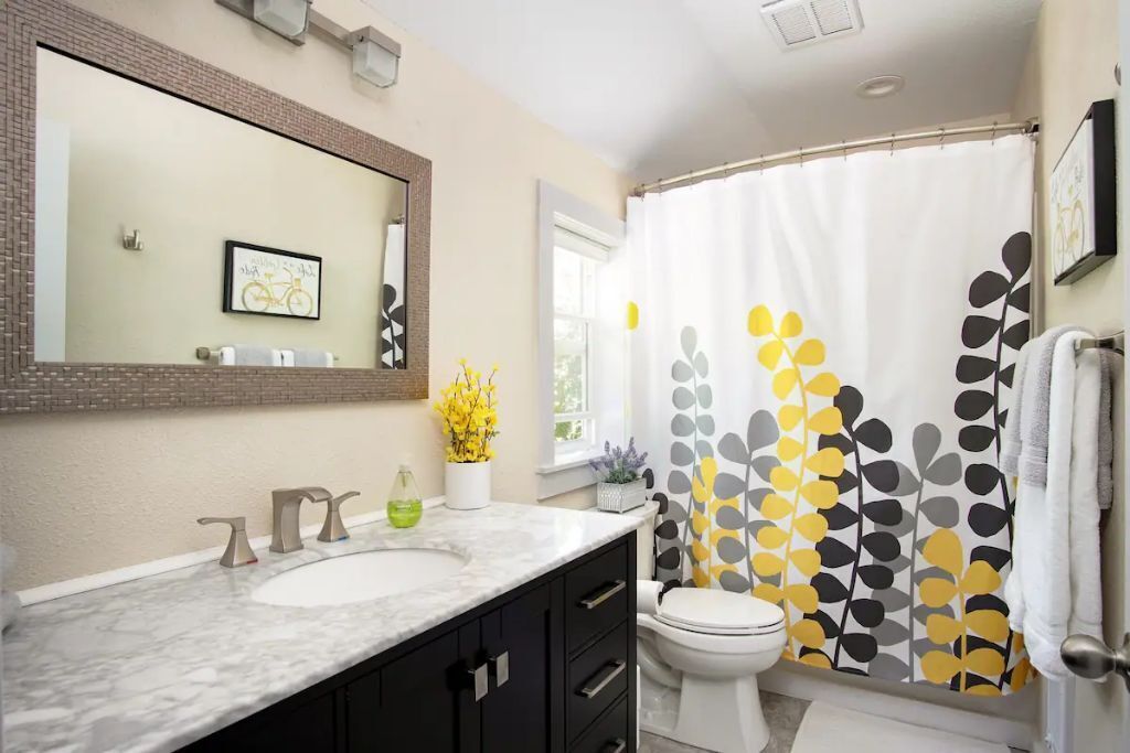 Une salle de bain avec un lavabo blanc, des meubles noirs, des toilettes blanches et un rideau de douche orné d’un motif de feuilles.