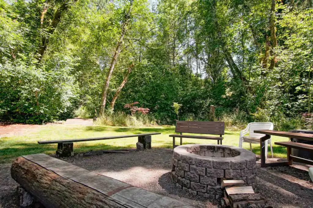 A forested outdoor area with a stone fire pit, benches, and picnic tables.