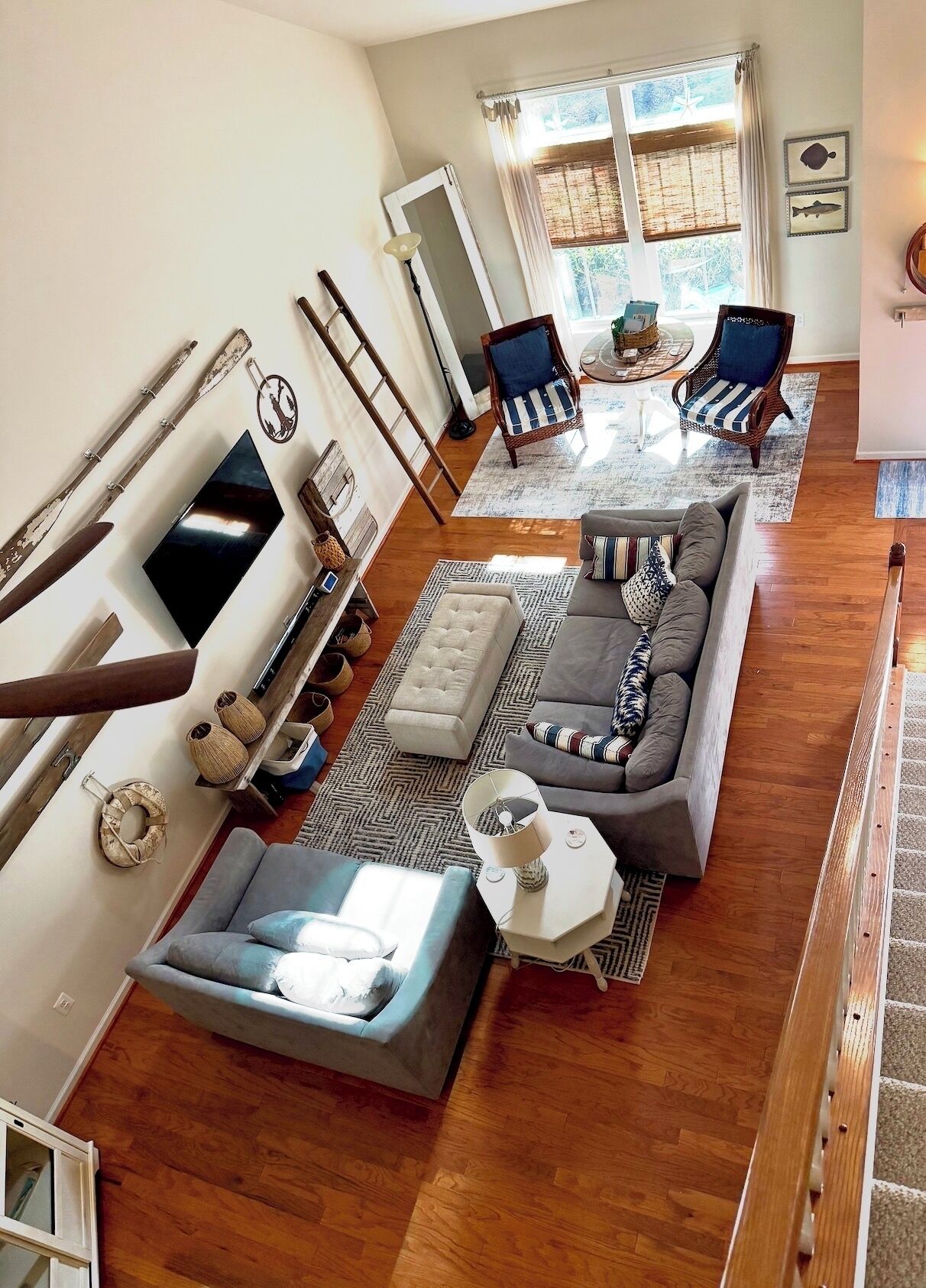 A spacious living room with a large gray sectional sofa, a small ottoman, a coffee table, and two armchairs. There is a wooden staircase on the right, a large window with curtains, and a wall-mounted TV.