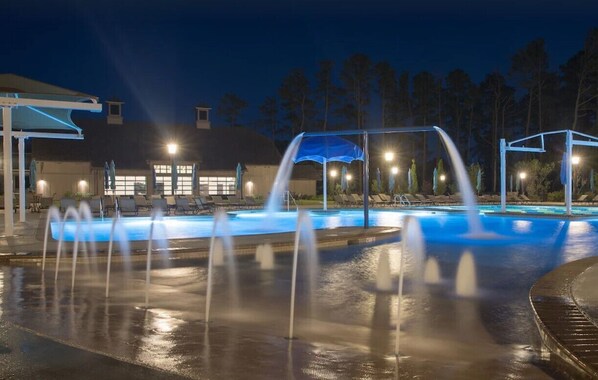 A swimming pool with multiple water jets at night, surrounded by umbrellas and lounge chairs.