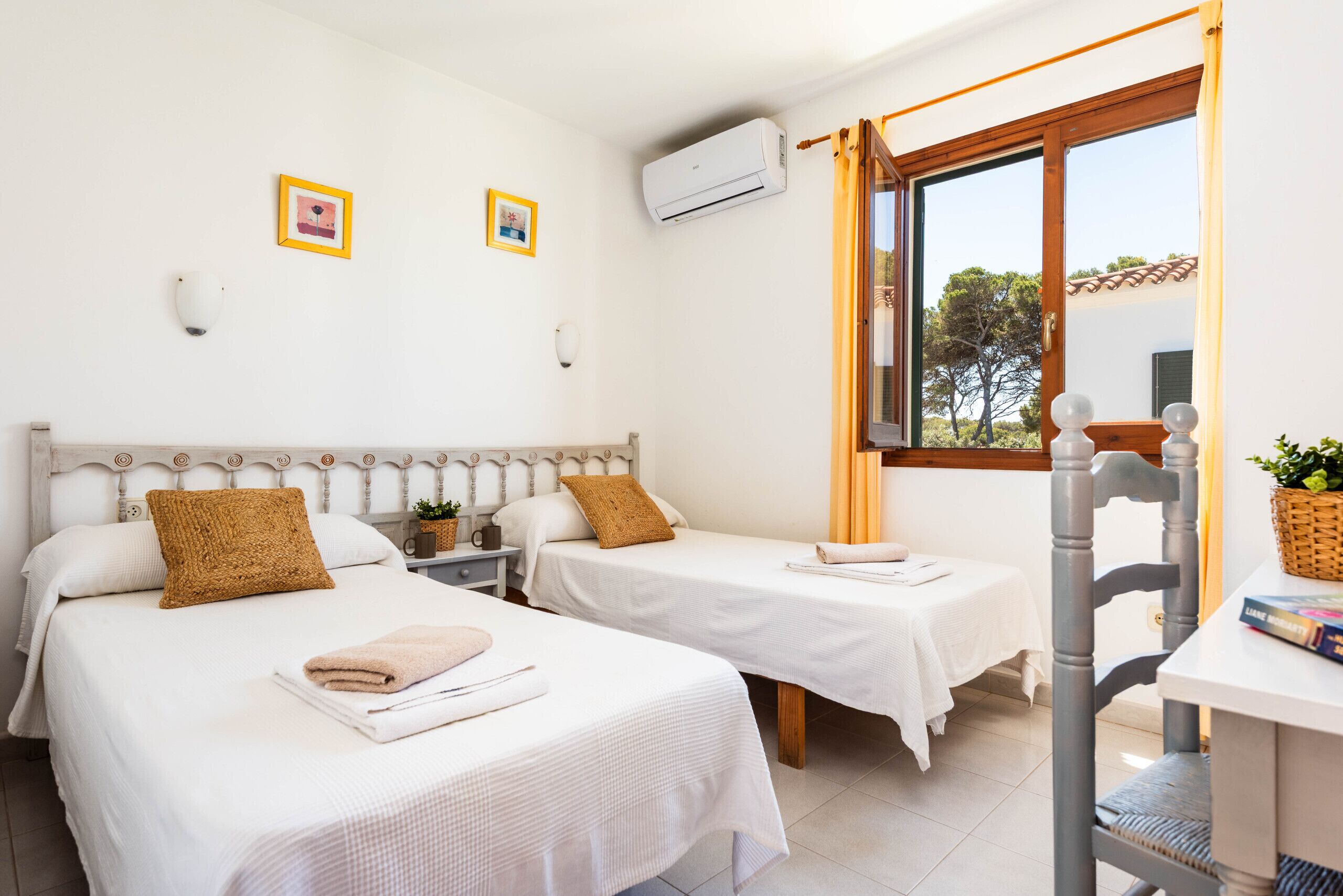 A room with two beds, white bedding, a wooden headboard, a small table with a basket, and a window with curtains.
